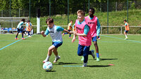 Zwei Kinder spielen Fußball auf einem Rasenplatz; ein Kind in Blau dribbelt den Ball, daneben ein Kind in Pink, im Hintergrund stehen weitere Kinder und ein Tor.