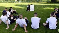 Fußballtrainer erklärt Taktik vor Gruppe von Spielern, die auf dem Rasen sitzen, mit Taktiktafel im Hintergrund.