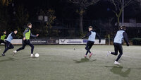 Spieler trainieren nachts auf einem Kunstrasenplatz, während ein Spieler mit dem Ball von Gegenspielern verfolgt wird.