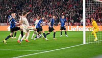 Fußballspiel mit mehreren Spielern, einer schießt auf das Tor, Torwart in Gelb bereitet sich auf den Schuss vor, im Hintergrund viele Fans.