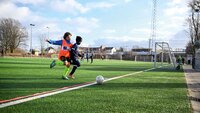 Zwei Kinder spielen Fußball auf einem Sportplatz, während andere im Hintergrund trainieren.