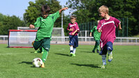 Kinder spielen Fußball auf dem Rasenplatz mit Hütchen und Minitor bei sonnigem Wetter.