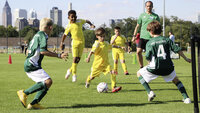 Kinder-Fußballspiel auf einer Wiese, Team in Gelb greift an gegen grün gekleidete Verteidiger vor kleinem Tor, mit Trainer am Rand und Skyline im Hintergrund bei Sonnenschein.