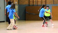 Kinder spielen Fußball in einer Turnhalle mit blauen Trikots und zwei Bällen.