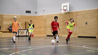Vier Kinder beim Fußballtraining in der Halle. Ein Junge im roten Trikot dribbelt, die anderen verfolgen ihn.