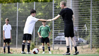 Trainer gibt einem Jugendspieler Feedback per Faustgruß auf dem Fußballplatz, Teamkollegen stehen im Hintergrund.