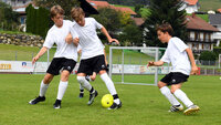 Kinder spielen Fußball auf einem grünen Rasenplatz in ländlicher Umgebung.