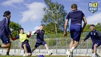 Fußballspieler in blauen Trainingsanzügen üben auf einem Sportplatz Zweikampfsituationen.