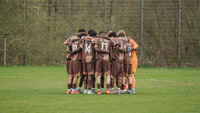 Eine junge Fußballmannschaft des FC St. Pauli in braunen Trikots schwört sich im Kreis auf dem grünen Rasen auf das Spiel ein.