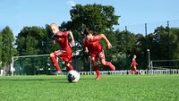 Zwei Kinder in roten Fußballtrikots spielen auf einem grünen Rasenplatz, eines dribbelt den Ball, im Hintergrund weitere Kinder und Bäume.