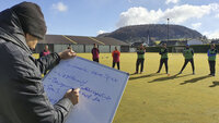 Trainer zeigt auf ein Whiteboard, während eine Gruppe Jugendlicher auf einem sonnigen Sportplatz zuhört; im Hintergrund Tore, Häuser und ein Hügel.