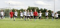 Kinder in Fußballtrikots laufen Hand in Hand über ein Spielfeld vor einem Tor.