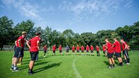 Gruppe von Fußballtrainern in roter Trainingskleidung steht im Kreis auf einem Fußballplatz und hört einem Vortrag zu.