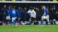 Ein Spieler im weißen Trikot dribbelt den Ball gegen zwei Spieler im blauen Trikot während eines Fußballspiels.