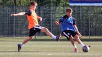Zwei jugendliche Fußballspieler im Training auf Kunstrasen: Ein Spieler im orangefarbenen Leibchen grätscht mit ausgestrecktem Bein, der andere im blauen Leibchen kontrolliert den Ball mit der Innenseite. Im Hintergrund ein hoher Zaun und Bäume, sonnige Bedingungen.