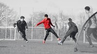 Ein Fußballspieler im roten Trikot dribbelt den Ball, während zwei Gegenspieler versuchen, ihn zu stoppen.