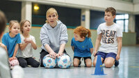 Kinder sitzen in einer Sporthalle im Kreis, zusammen mit einer erwachsenen Betreuungsperson; vor ihnen liegen Bälle und ein blauer Kegel.