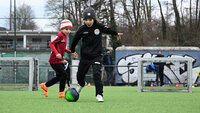 Zwei Kinder beim Fußballtraining auf einem Kunstrasenplatz; eines im schwarzen Trainingsoutfit dribbelt einen Ball, eine weiteres in roter Jacke läuft daneben.