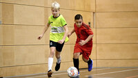 Zwei Kinder spielen in einer Sporthalle Fußball und rennen dem Ball hinterher.