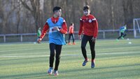 Zwei junge Fußballspieler trainieren auf einem Sportplatz, umgeben von weiteren Spielern im Hintergrund.