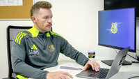 Fußballtrainer sitzt am Schreibtisch und schaut auf einen Computerbildschirm mit Vereinslogo.