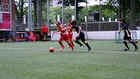 Kinder spielen Fußball auf einem kleinen Kunstrasenplatz, Spieler in Rot und Blau kämpfen um den Ball.