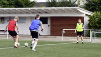 Drei Fußballer im Training auf Kunstrasen, ein Pass vom roten zum blauen Spieler, der gelbe verteidigt vor einer kleinen Tribüne.