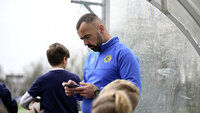 Ein Fußballtrainer im blauen Trainingsanzug schaut am Spielfeldrand konzentriert auf sein Smartphone.