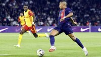 Kylian Mbappé von Paris Saint-Germain im blauen Trikot dribbelt dynamisch an einem Gegenspieler im gelb-roten Trikot vorbei.