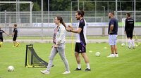 Personen stehen und unterhalten sich am Spielfeldrand eines Fußballtrainings, während Kinder im Hintergrund trainieren.