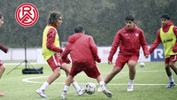 Trainingsspiel bei Regen: Spieler in Rot lösen eine enge Pressingsituation im Zentrum, RWE-Logo eingeblendet.
