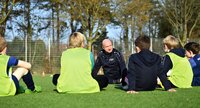 Ein Jugendtrainer spricht auf dem Fußballplatz zu seiner Mannschaft, die ihm im Kreis gegenübersitzt.