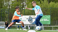 Zwei Kinder duellieren sich im Fußball: ein Kind in Orange grätscht, ein Kind in Blau-Weiß versucht am Ball vorbeizuziehen, im Hintergrund Werbebanden und Bäume.