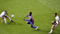 Ein Spieler in einem blau-roten Trikot dribbelt den Ball zwischen zwei Verteidigern in weißen Trikots.