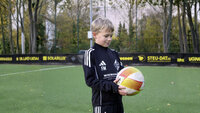 Junge im schwarzen Trainingsanzug hält Fußball auf Kunstrasenplatz, Werbebanden und Bäume im Hintergrund.