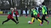 Kinder spielen Fußball auf einem Trainingsplatz, Zuschauer stehen am Rand.