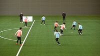 Jugendliche spielen Fußball in einer Halle auf einem Kunstrasenfeld.