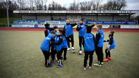 Zwei Trainer geben neun Kindern im blauen Trainingsanzug Anweisungen beim Fußballtraining.