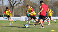 Jugendliche spielen Fußballtraining auf einem Rasenplatz; ein Spieler im roten Shirt dribbelt den Ball zwischen gelben Markierungshütchen, weitere Mitspieler in gelben Leibchen üben im Hintergrund.