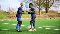 Zwei Kinder spielen mit einem Ballon während eines Fußballtrainings auf einem Kunstrasenplatz.