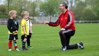 Ein Trainer spricht kniend mit drei Kindern auf dem Sportplatz und gibt ruhige Anweisungen.
