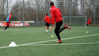 Fußballtraining auf Kunstrasen: mehrere Spieler in roten Trikots sprinten zwischen Markierungshütchen; im Hintergrund Tore und Bäume bei grauem Wetter.