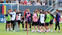 Eine Fußballmannschaft steht in einem Kreis auf einem Trainingsplatz, während ein Trainer Anweisungen gibt.