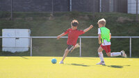 Zwei Kinder auf dem Fußballplatz, eines bereitet sich darauf vor, den Ball zu schießen, Trainingsplatzabgrenzung im Hintergrund.