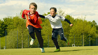 Zwei Kinder rennen auf einem Rasenfeld; links ein Kind im roten Trikot, rechts ein Kind im hellen Oberteil, beide in Bewegung vor grünem Hintergrund.