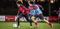 Fußballspieler trainieren auf einem beleuchteten Platz, ein Spieler dribbelt den Ball unter Druck eines Gegenspielers.