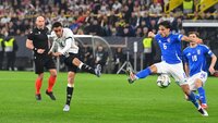 Ein Spieler in weißem Trikot schießt den Ball, während ein Spieler in blauem Trikot versucht, den Schuss zu blocken.