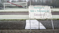 Ein Schild "Rasenplatz gesperrt" steht im Winter vor einem vereisten Fußballplatz und signalisiert die Winterpause.