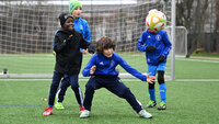 Drei Kinder in blauen Trainingssachen spielen Fußball auf einem Kunstrasenplatz vor einem Tor; eines schießt oder passt den Ball, zwei weitere beobachten und reagieren.
