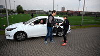 Vater übergibt seinem Sohn die Trainingstasche, Mutter sitzt lächelnd im weißen Auto, im Hintergrund ein Fußballtrainingsplatz.